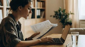 Une jeune femme assise à une table remplit sa demande AAH sur un ordinateur portable, avec un dossier papier et un stylo à côté d'elle.